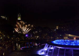 A pyrotechnic show in front of Downtown Los Angeles' City Hall as Gloria Molina Grand Park celebrates the 4th of July Block Party. A fountain is lit with colors illuminating the night atmosphere in the park.